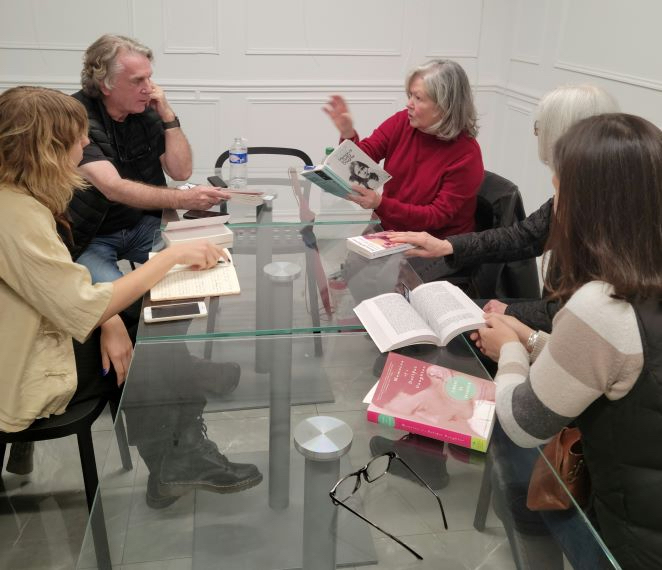 Photo of a book club. Four older white women and a man are sitting around a glass table discussing a book.