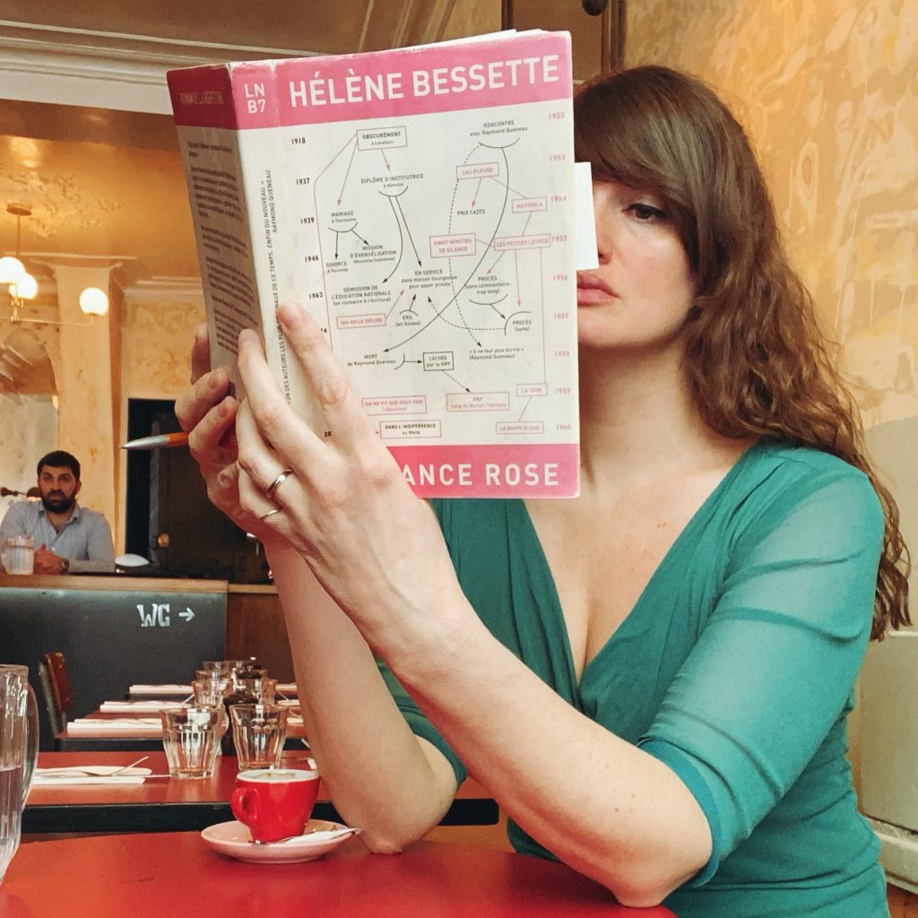 Carrie Chappell sits at a cafe table, half hidden behind her book Garance Rose by Helene Bessette